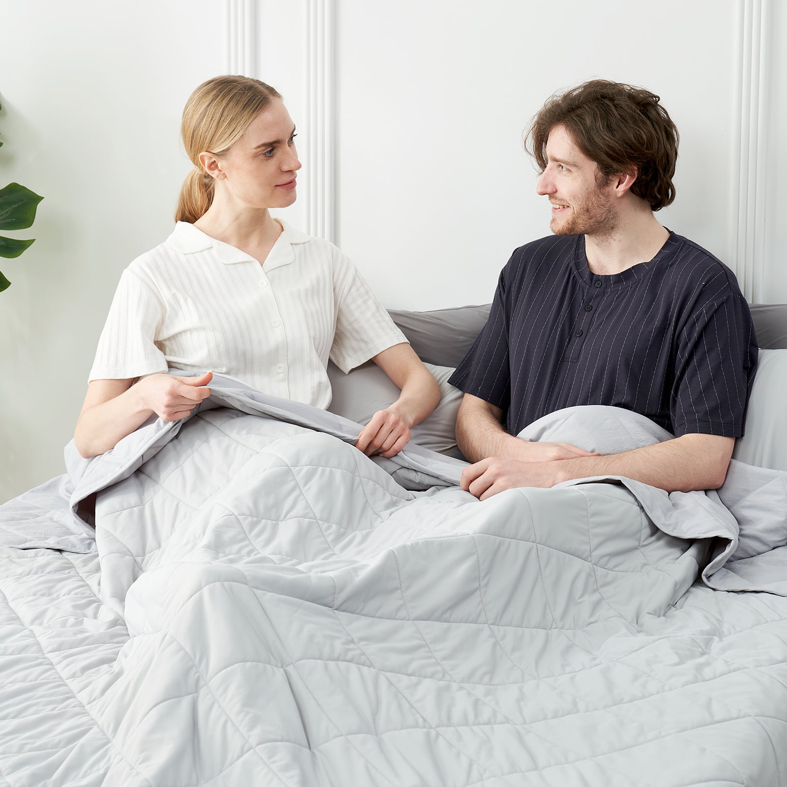 Couple sitting on a bed with a grey cooling comforter, surrounded by a minimalistic room.#color_Light Grey