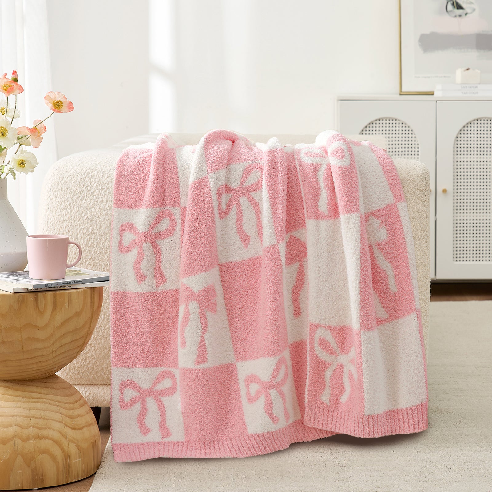 Snuggle Sacs Reversible Checkered Bow Feather Yarn Knit Throw Blanket on a beige couch with mug and a vase of flower#color_Pink_Bow