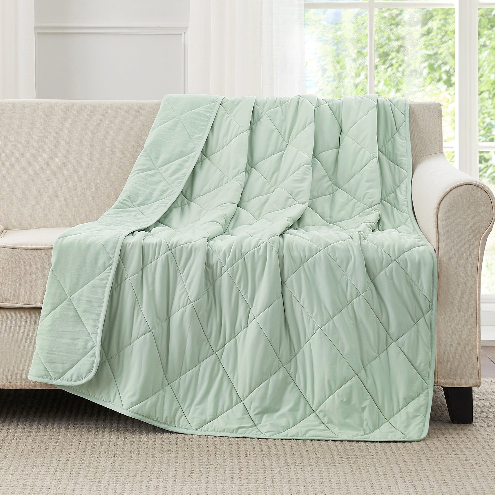 Snuggle Sac Green cooling blanket draped over a beige sofa in a room with large windows.#color_Mint Green