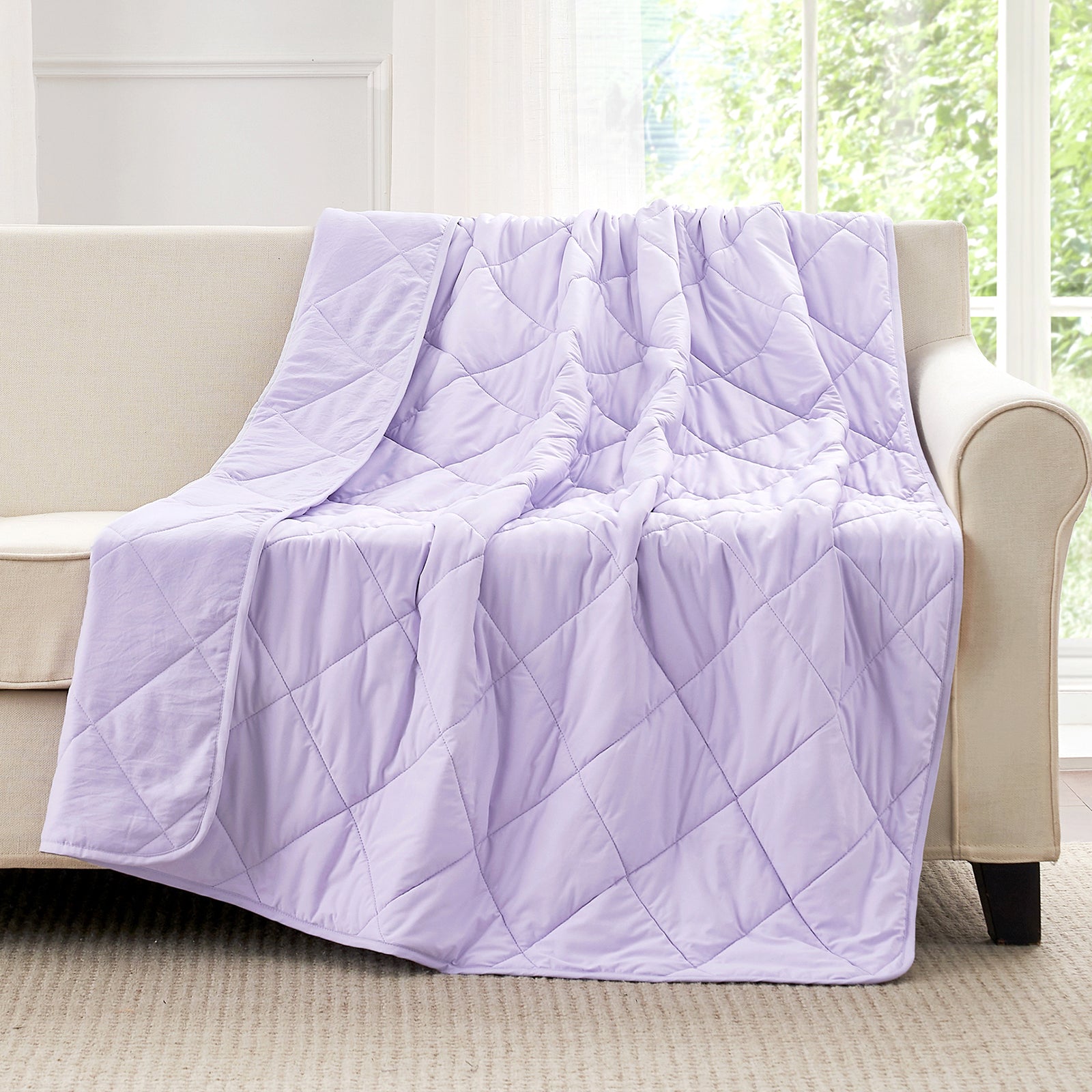 Snuggle Sac Purple cooling blanket draped over a beige sofa in a room with large windows.#color_Purple