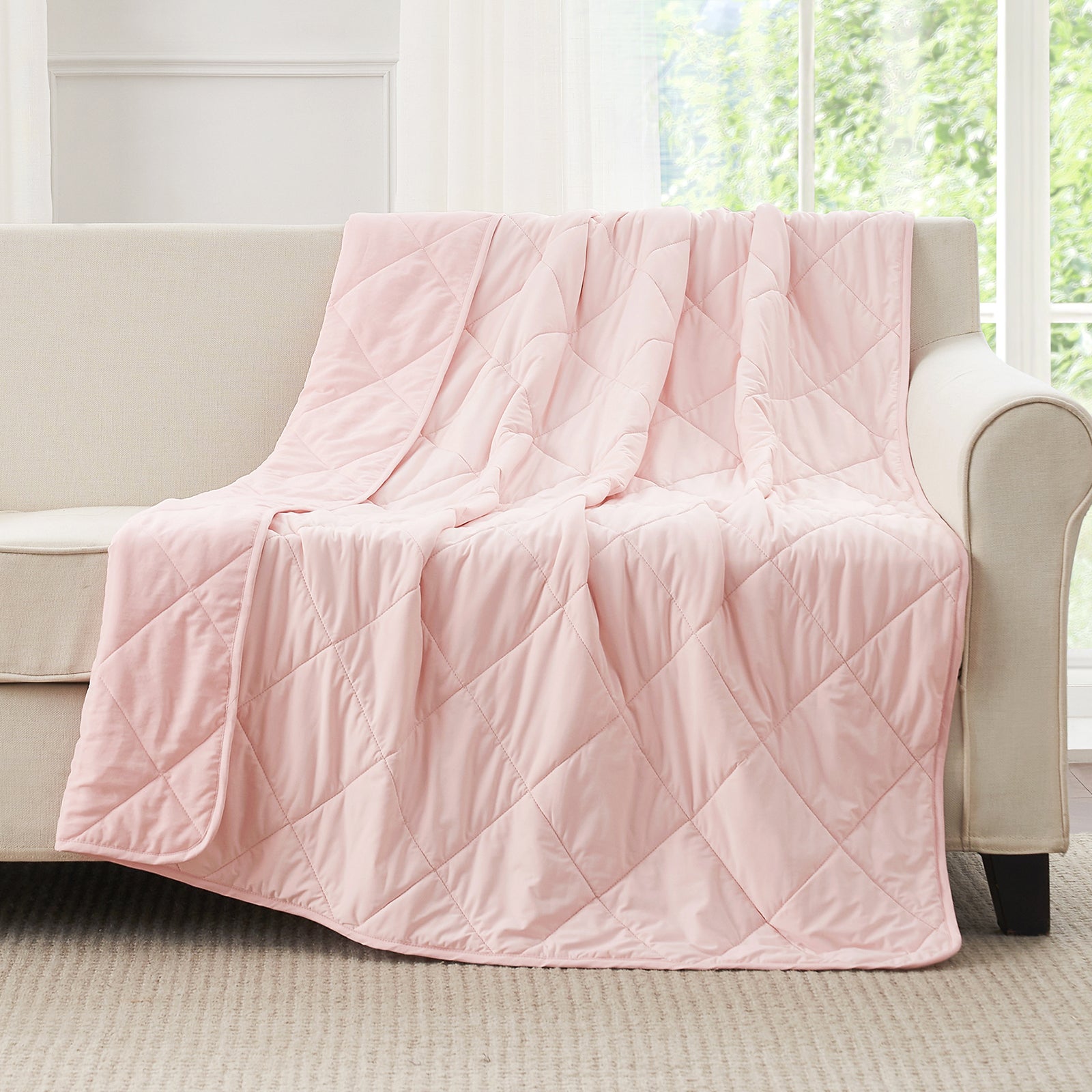 Snuggle Sac Pink cooling blanket draped over a beige sofa in a room with large windows.#color_Pink