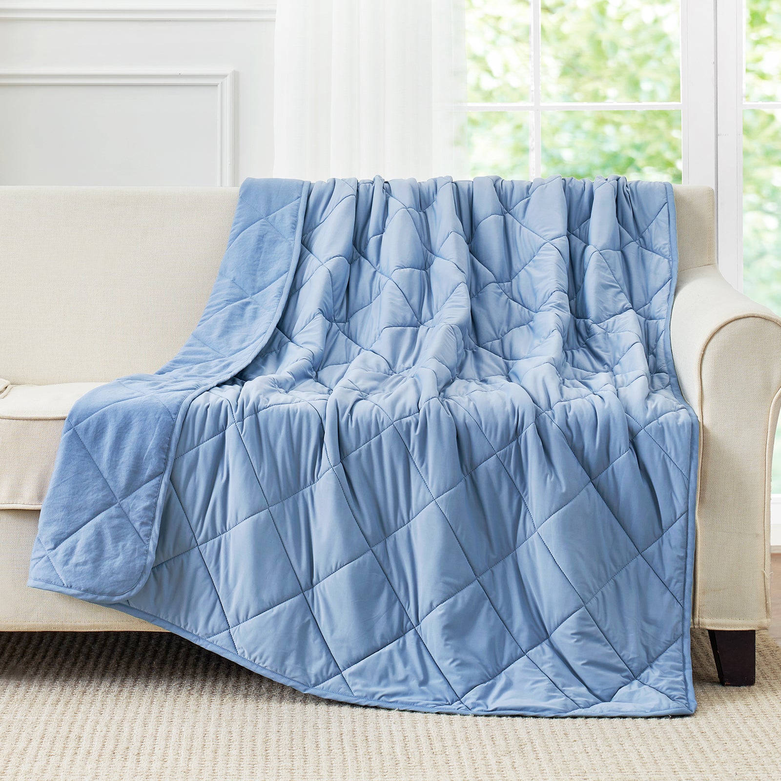 Blue cooling blanket draped over a beige sofa in a room with large windows.#color_Dark Blue