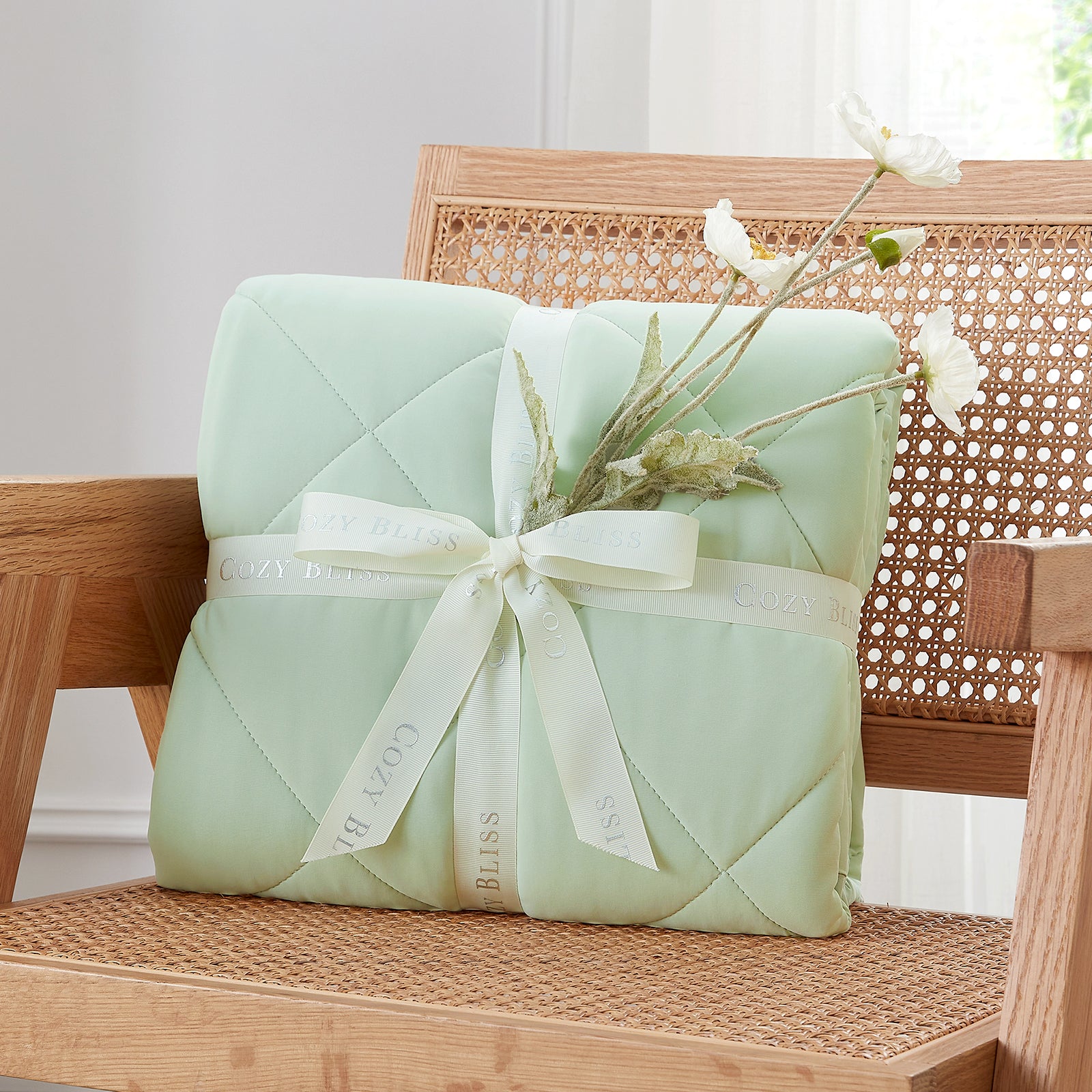 Folded Snuggle Sac mint green comforter with white ribbon on chair surrounded by plants, presented as a perfect gift#color_Mint Green