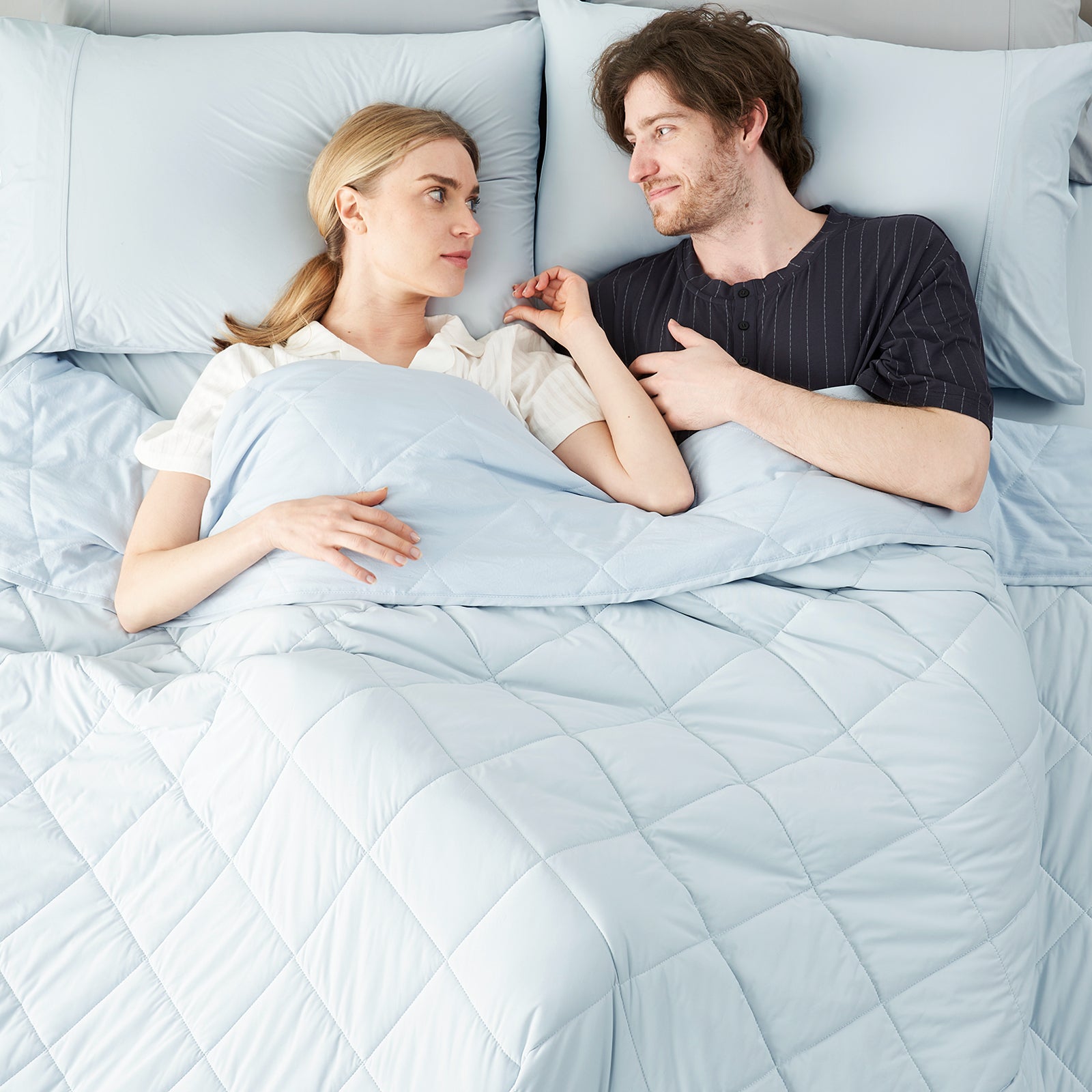 Couple lying on bed with a Snuggle Sac blue cooling comforter, surrounded by a minimalistic room.#color_Blue