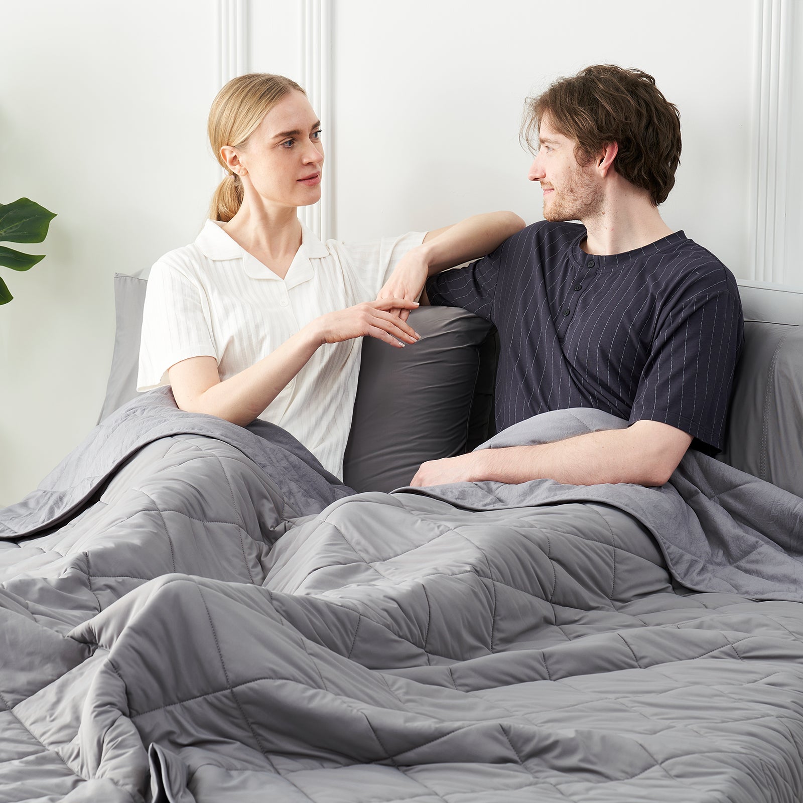 Couple sitting on a bed with a Snuggle Sac dark grey cooling comforter, surrounded by a minimalistic room.#color_Dark Grey