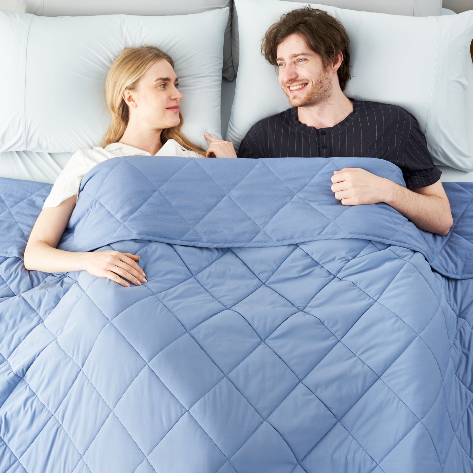 Couple lying on bed with a Snuggle Sac dark blue cooling comforter, surrounded by a minimalistic room.#color_Dark Blue