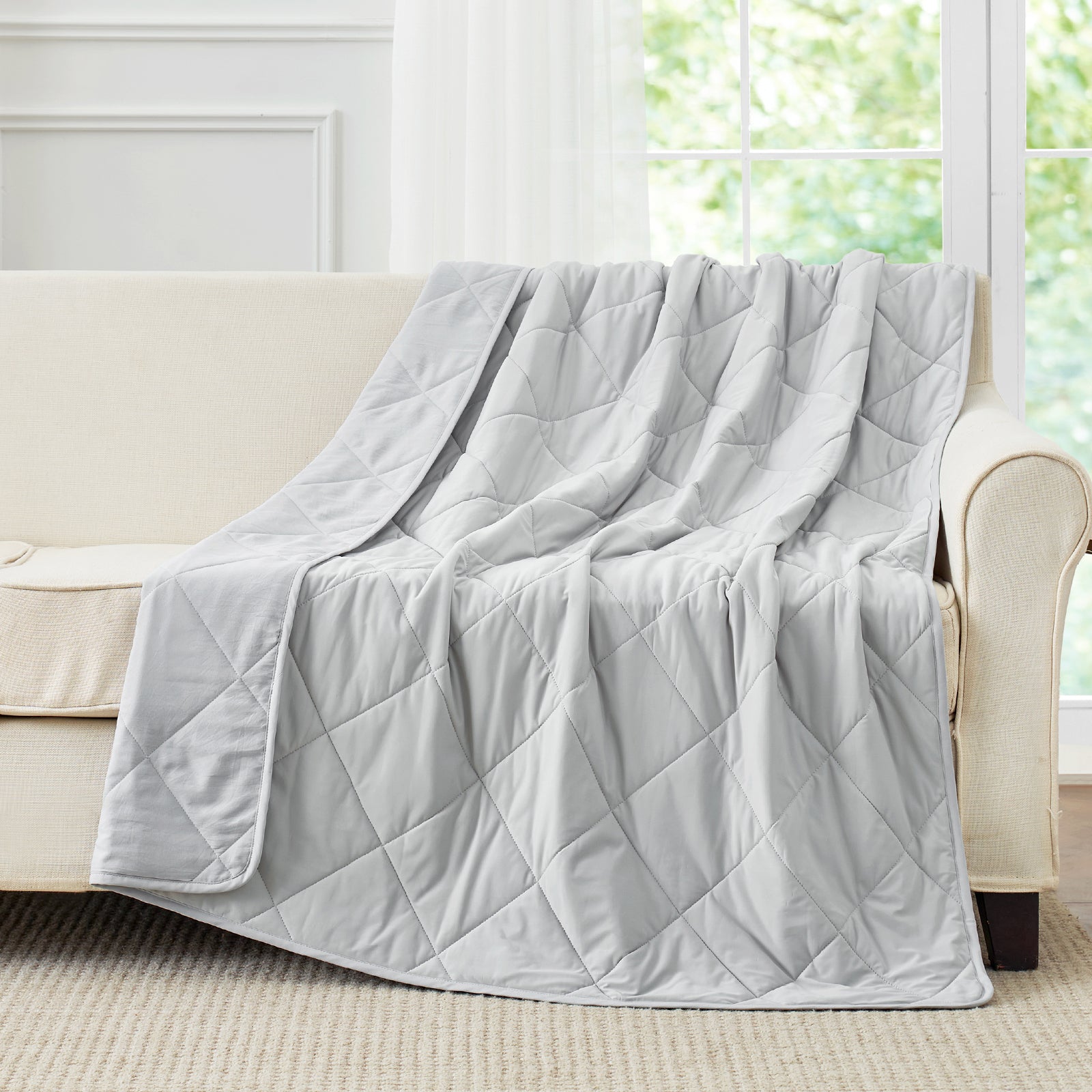 Snuggle Sac Gray cooling blanket draped over a beige sofa in a room with large windows.#color_Light Grey