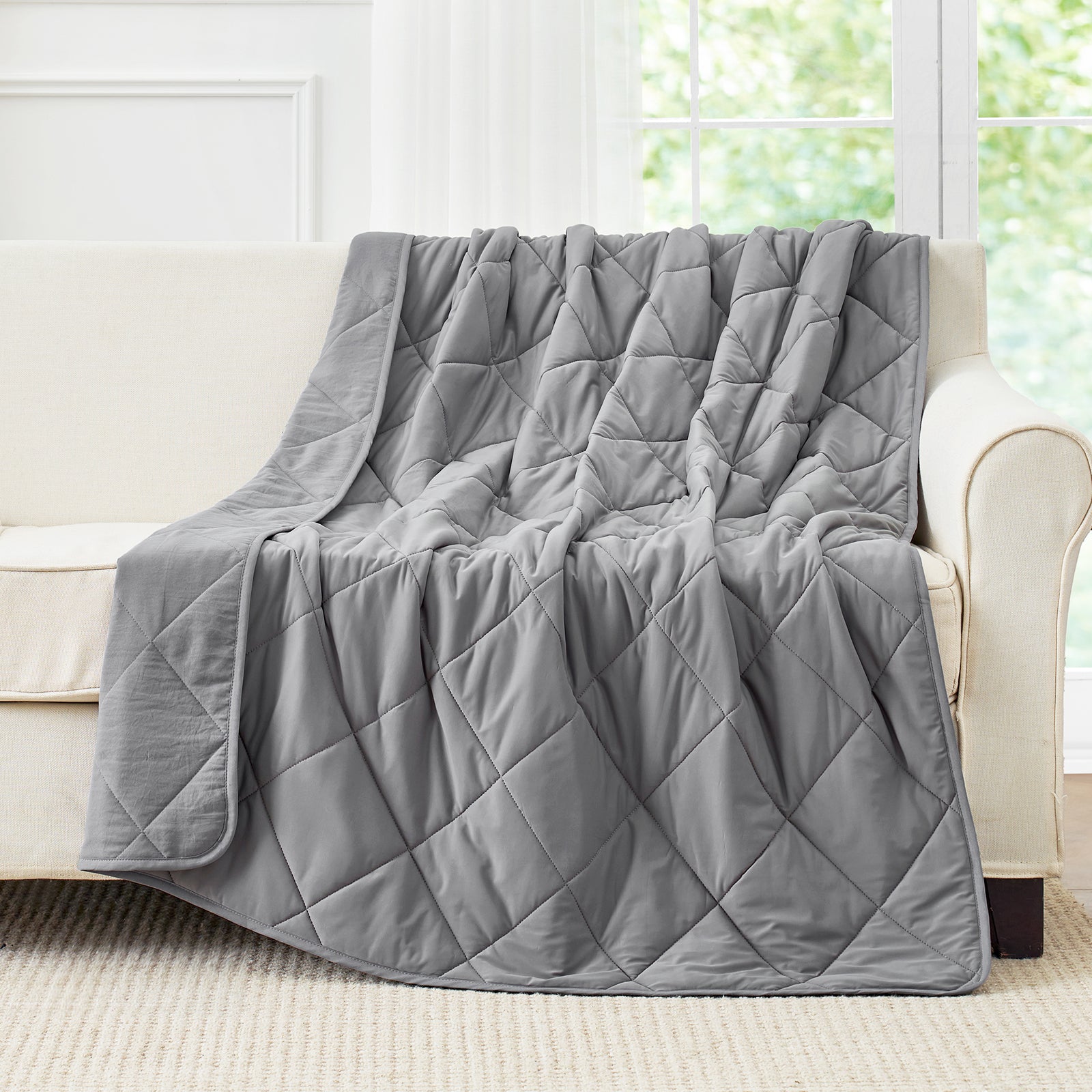 Snuggle Sac Dark Grey cooling blanket draped over a beige sofa in a room with large windows.#color_Dark Grey