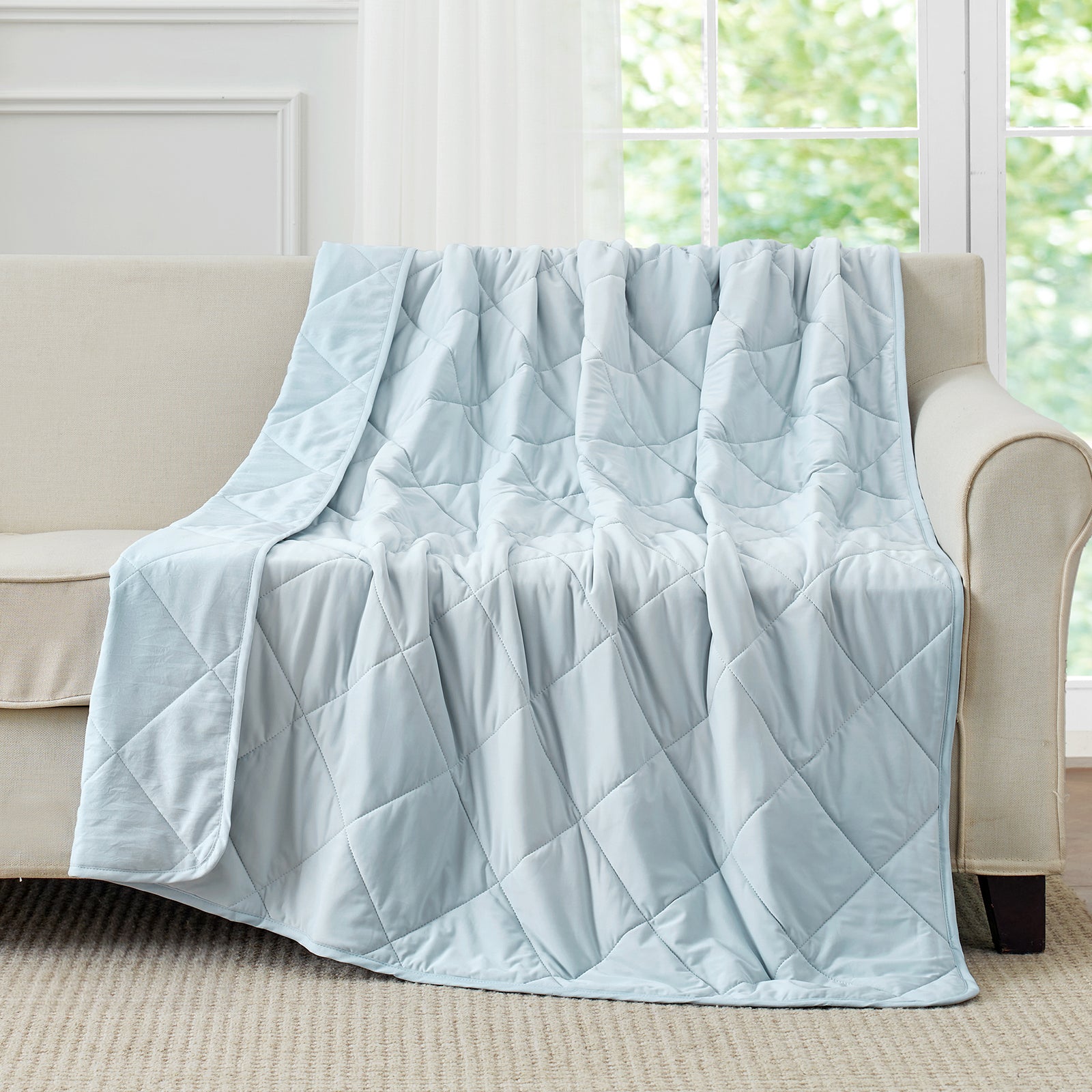 Snuggle Sac Blue cooling blanket draped over a beige sofa in a room with large windows.#color_Blue
