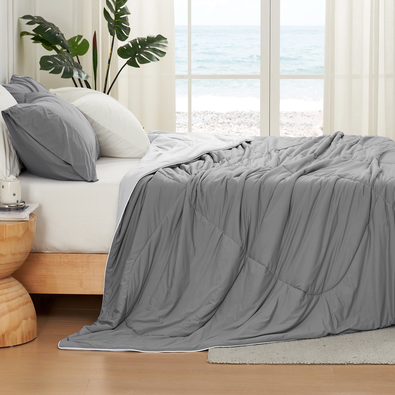 Left side view of grey Snuggle Sac comforter on a bed with a window view of the ocean#color_Dark Grey
