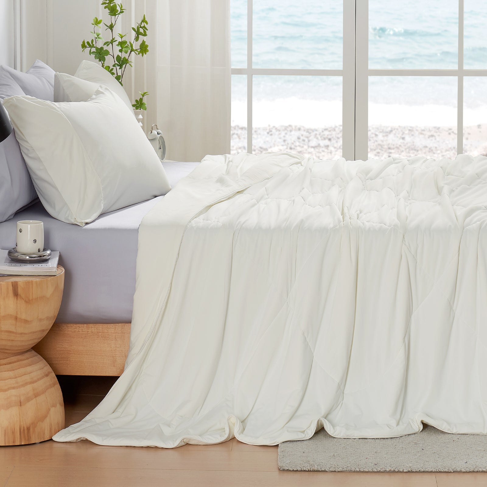 Left side view of Snuggle Sac cream white cooling comforter on a bed in a bright bedroom with large windows.#color_Cream White