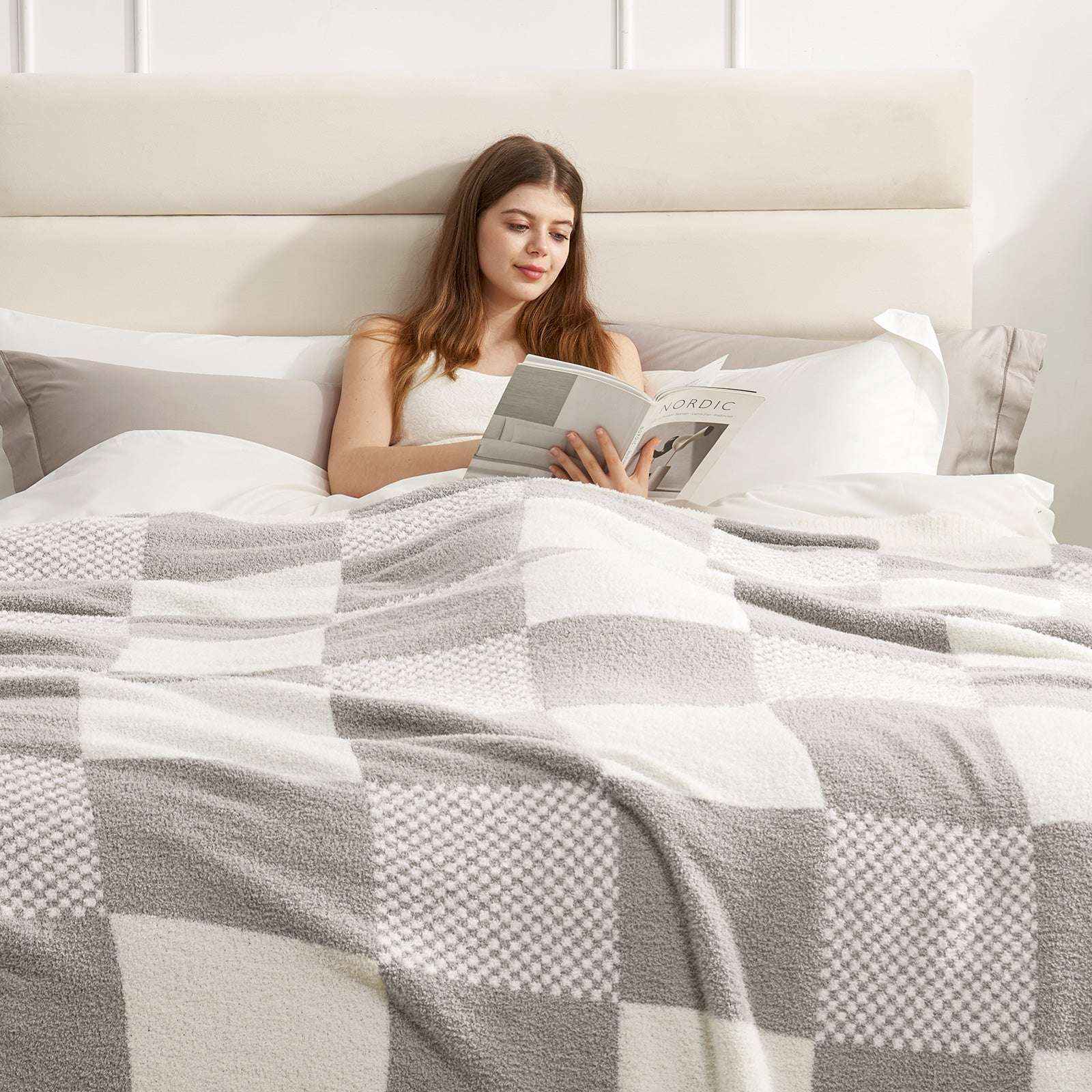 A woman laying on bed reading a book with Snuggle Sac Checkered Feather Yarn Knit Throw Blanket on her#Color_Grey