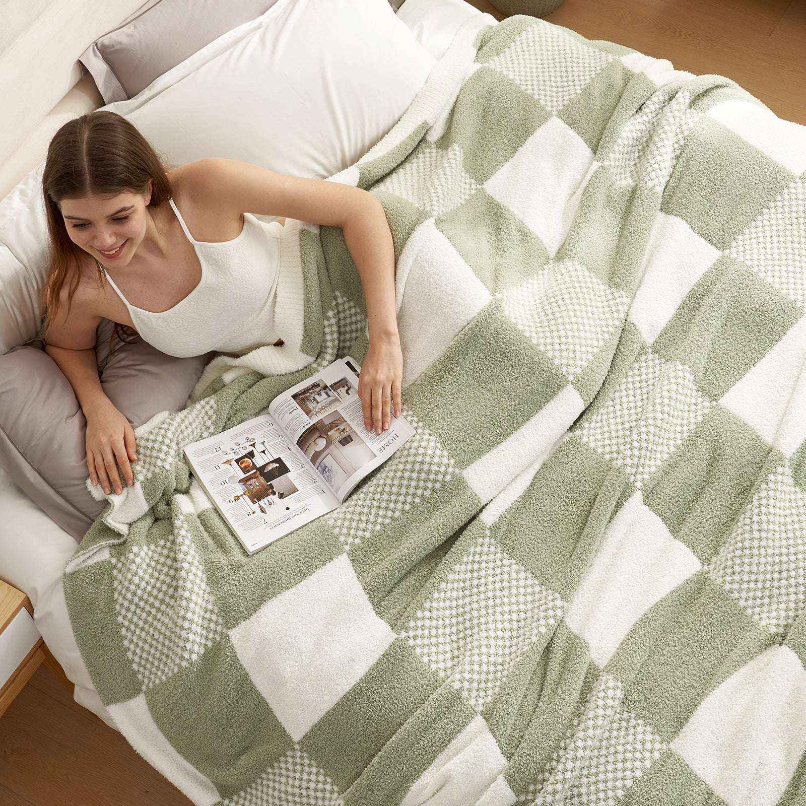 A woman laying on bed reading a book with Snuggle Sac Checkered Feather Yarn Knit Throw Blanket on her#Color_Sage Green
