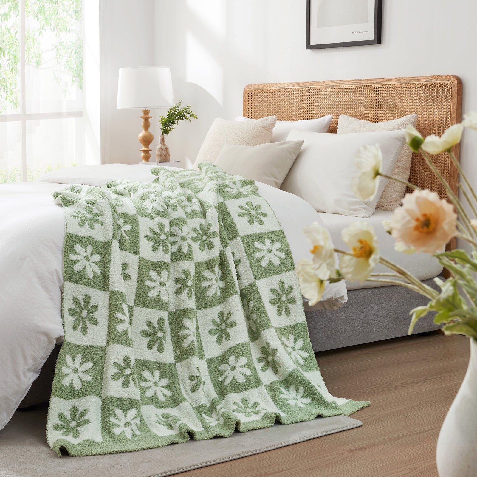 Green and white patterned blanket on a bed in a bright bedroom with flowers and a lamp.  #color_Sage Green