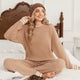A woman sits cross-legged on a white bed wearing a cozy knit lounge set in a warm taupe-brown color, consisting of a long-sleeve sweater and matching pants. She is also wearing a matching knit beanie.#color_Khaki