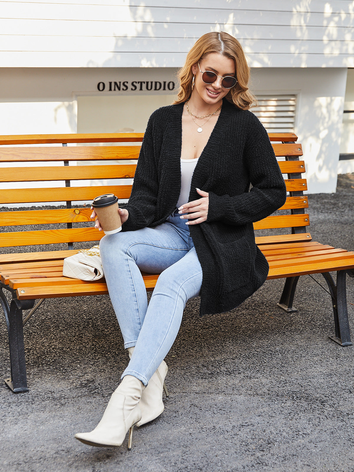 Woman sitting on a bench wearing a black cardigan, light blue jeans, and white boots, holding a coffee cup. #color_Black 