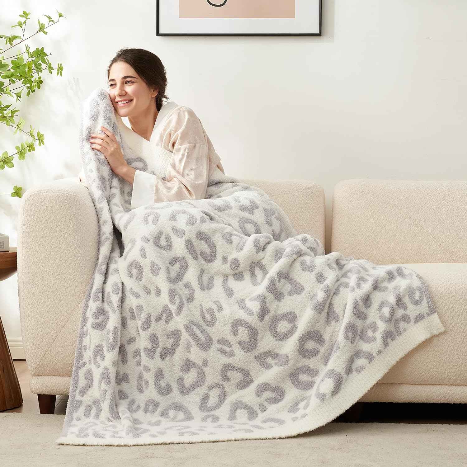 A woman sitting on a beige couch wrapped in Reversible Leopard Feather Yarn Knit Throw Blanket#Color_Leopard Grey