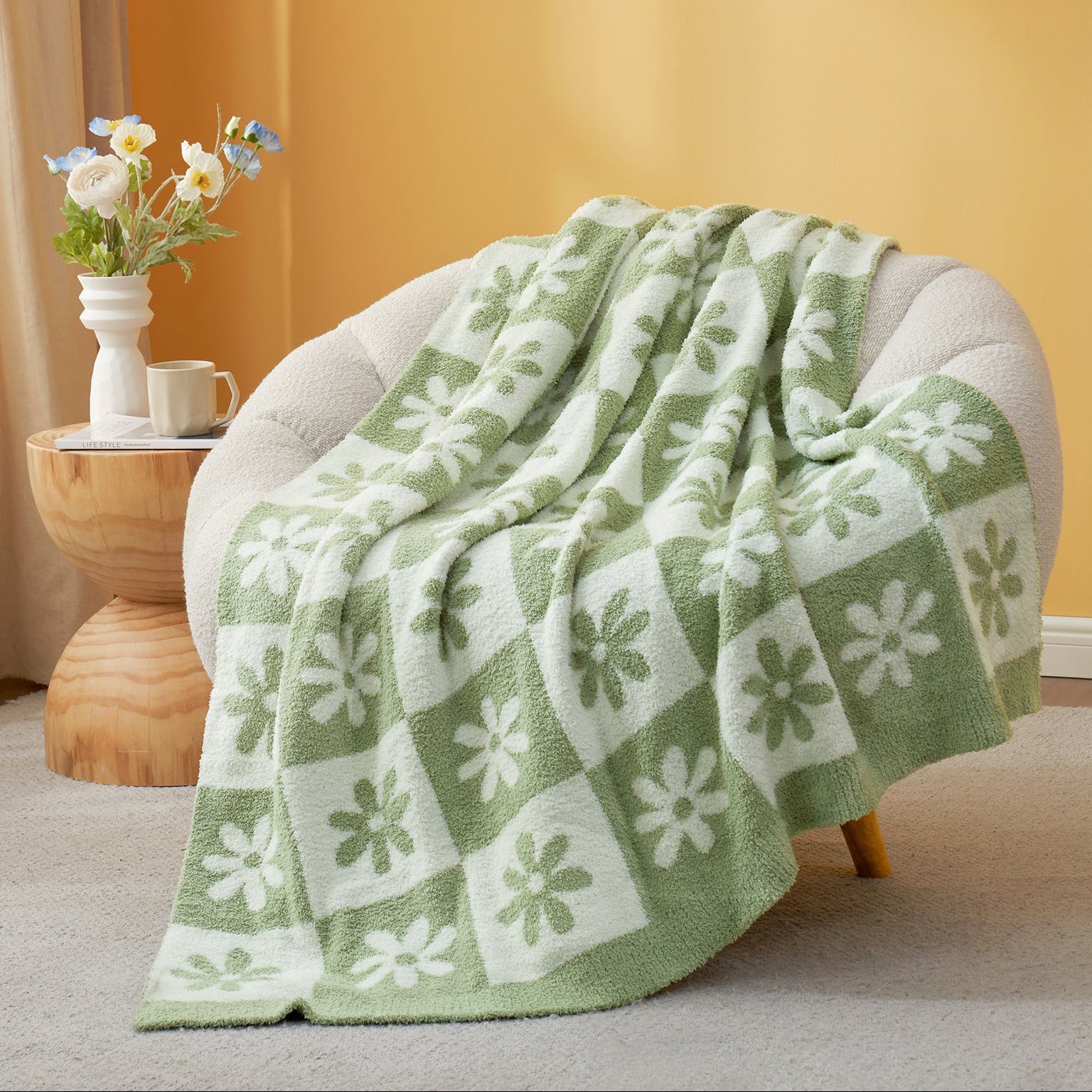 Green and white patterned blanket draped over a chair with a side table and flowers in the background.  #color_Sage Green