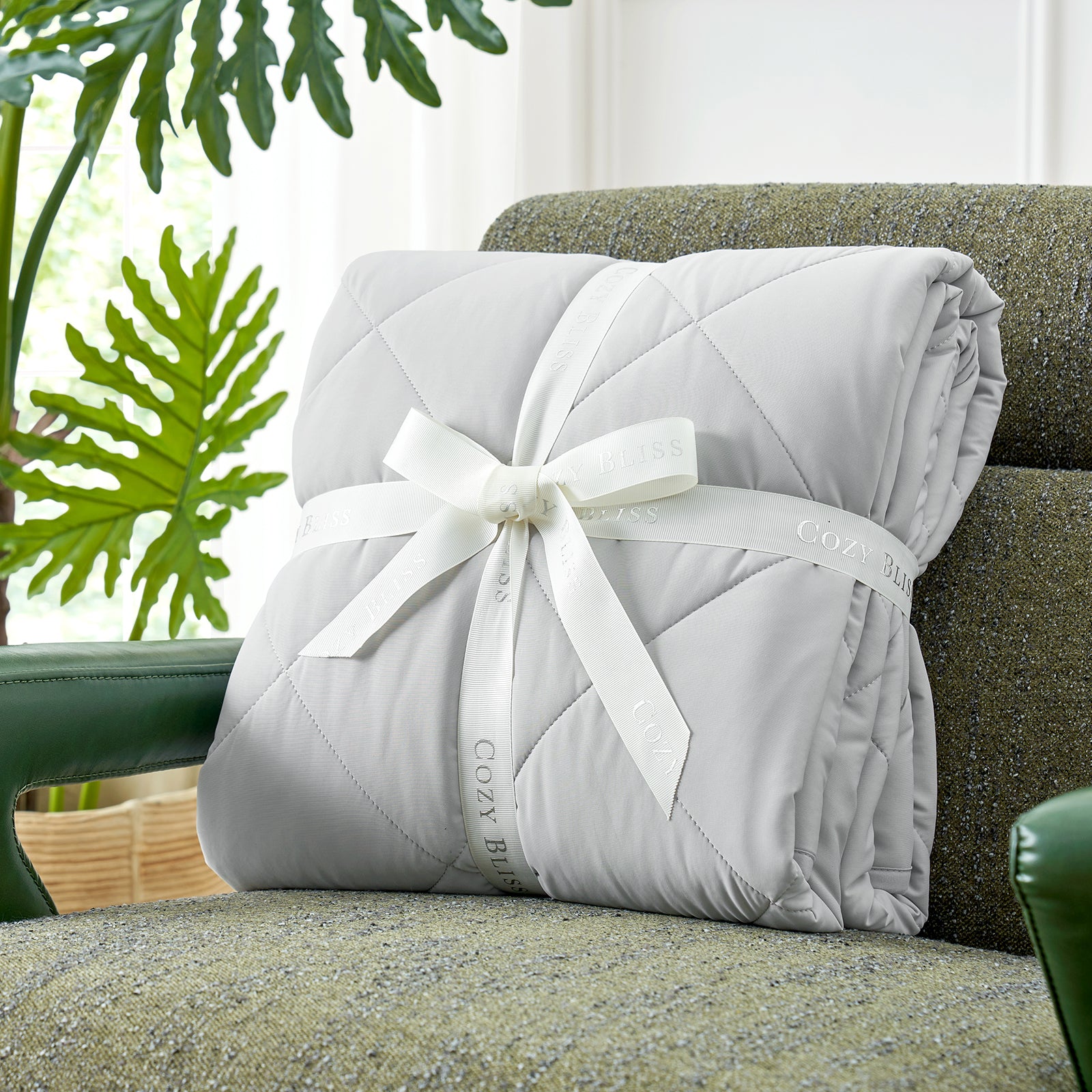 Folded comforter with white ribbon on green couch surrounded by plants, presented as a perfect gift#color_Light Grey