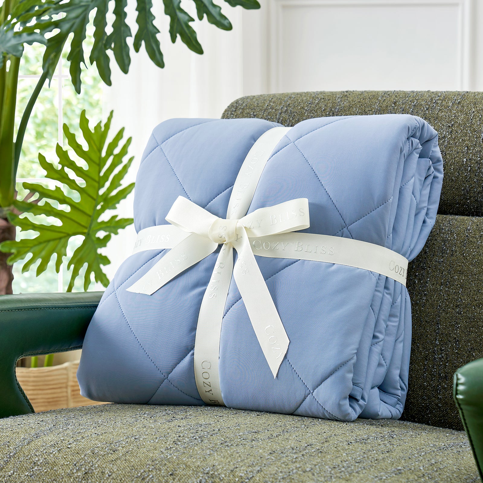 Folded comforter with white ribbon on green couch surrounded by plants, presented as a perfect gift#color_Dark Blue