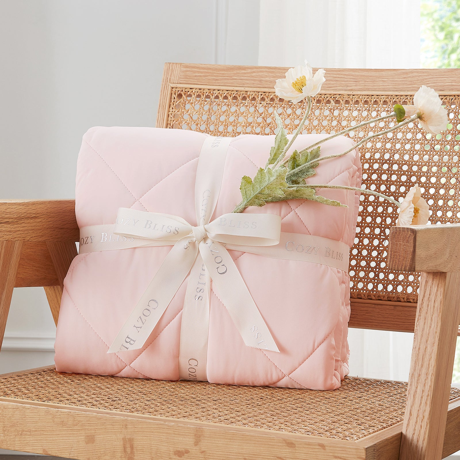 Folded Snuggle Sac pink comforter with white ribbon on chair surrounded by plants, presented as a perfect gift#color_Pink