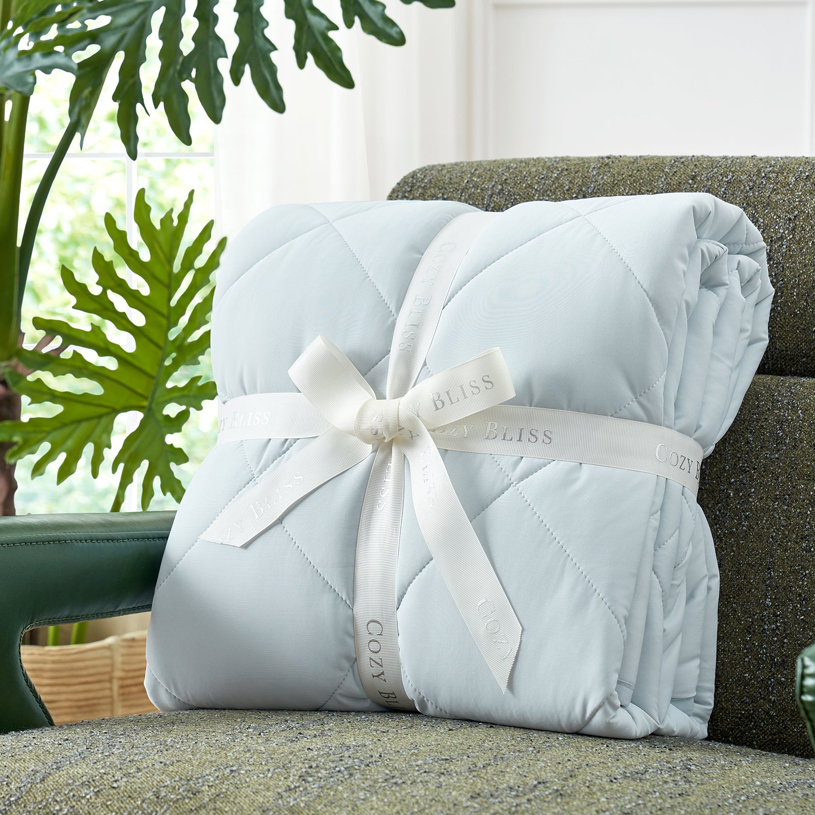 Folded Snuggle Sac comforter with white ribbon on green couch surrounded by plants, presented as a perfect gift#color_Blue