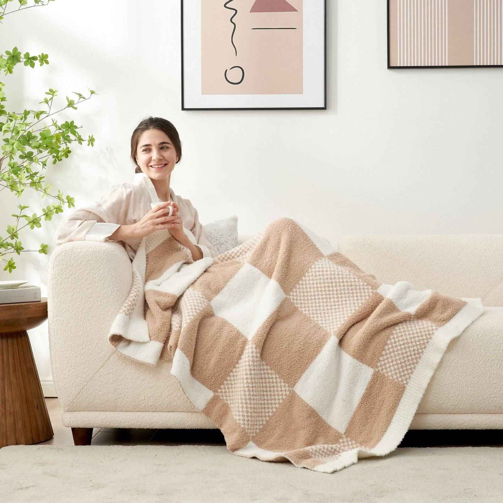 A woman laying down on the sofa reading a book with Snuggle Sac Checkered Feather Yarn Knit Throw Blanket on her lap#Color_Beige