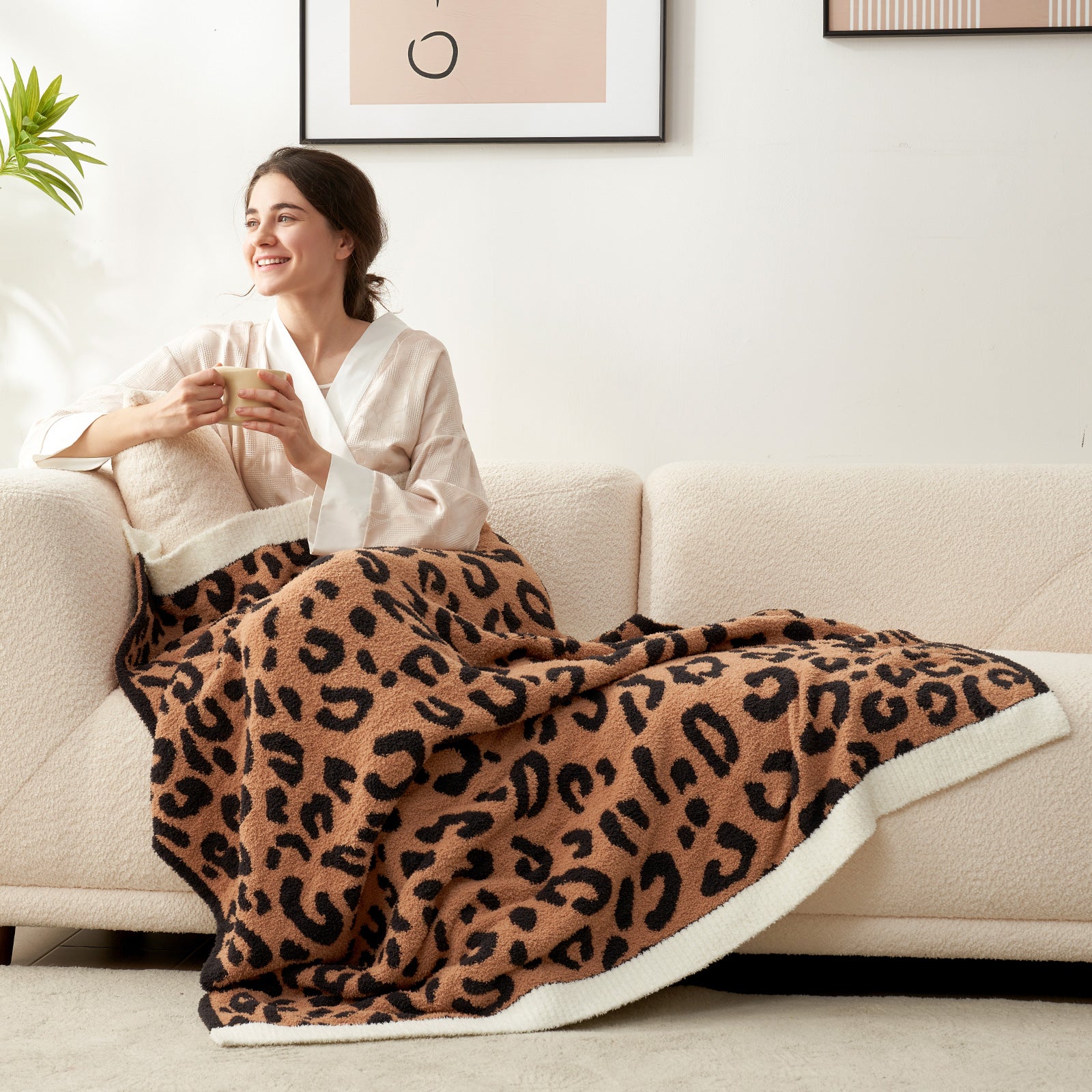 A woman sitting on a beige couch wrapped in Reversible Leopard Feather Yarn Knit Throw Blanket with a mug #Color_Black