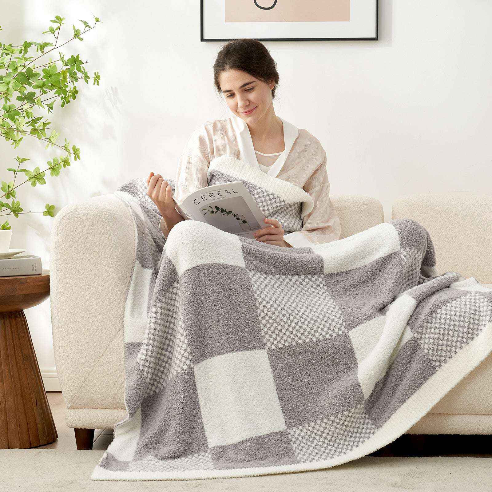 A woman sitting down on the sofa reading a book with Snuggle Sac Checkered Feather Yarn Knit Throw Blanket on her lap#Color_Grey